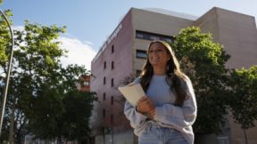 Une étudiante marche, souriante, avec des documents d'inscription dans les bras, devant l'université.