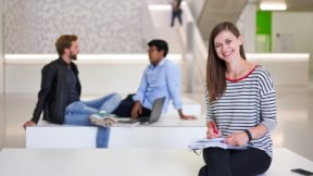 Une étudiante est assise, souriante, avec un bloc-notes dans le foyer de l'université.